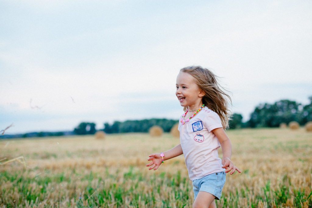a little girl wearing a pink shirt and light blue shorts runs through a cropped hay field